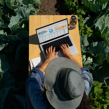 Vue en plongée d’une femme coiffée d’un chapeau à larges bords et installée à une table de jardin, préparant une présentation d’affaires sur un MacBook.