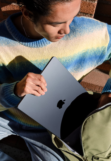 Un étudiant assis dehors. Il sort un MacBook fermé de son sac à dos.